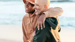 Two men are walking along the beach on a sunny day. One man has his arm around the other. They are both smiling.