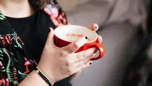 Close-up of a woman's hands with black-painted nails holding a red cup of tea