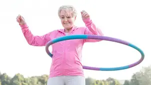 A woman uses a hula hoop outdoors, dressed in a pink hoodie and grey trousers